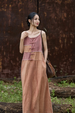 A woman wearing a long, sleeveless, two-tone dusty rose and brown maxi dress with traditional Chinese-style buttons, standing outdoors in front of a rusty metal wall.